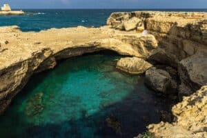 Ein großer natürlicher Felsbogen spannt sich über türkisfarbenes, klares Wasser in einer Küstenbucht. Zerklüftete Kalksteinfelsen umgeben den Pool, während im Hintergrund das offene Meer und die Wellen unter einem teilweise bewölkten Himmel zu sehen sind.