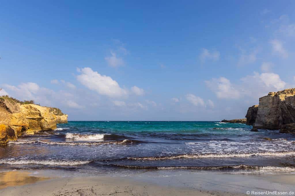 Ein Sandstrand mit sanften Wellen rollt vom türkisfarbenen Meer unter einem blauen Himmel mit vereinzelten weißen Wolken heran. Felsige Klippen mit spärlicher Vegetation umrahmen die Küstenlinie auf beiden Seiten. Die Szene wirkt ruhig und einladend.