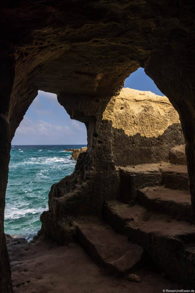 Blick aus dem Inneren einer Felsenhöhle mit grob behauenen Steinstufen, die zu einer unregelmäßigen Öffnung hinaufführen. Das Sonnenlicht dringt durch den Eingang und beleuchtet die strukturierten Felswände. Draußen schlagen türkisfarbene Wellen unter einem blauen Himmel mit vereinzelten Wolken gegen das Ufer.