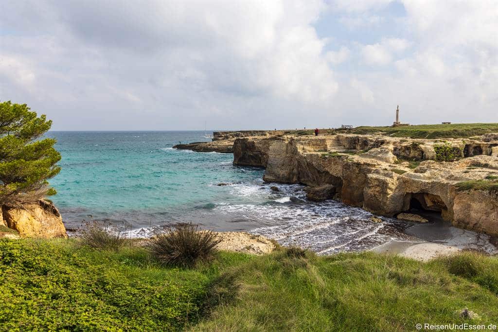Eine felsige Küste mit türkisfarbenen Wellen, die an eine kleine Sandbucht gespült werden, die von Klippen und grasbewachsenem Land umgeben ist. Auf der linken Seite steht ein einzelner Baum, und in der Ferne ist bei teilweise bewölktem Himmel ein Leuchtturm zu sehen.