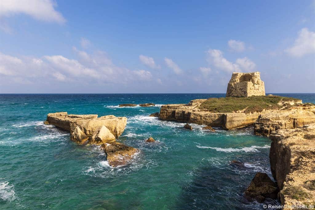 Ein verfallener Steinturm steht auf einer felsigen Landzunge, die ins türkisfarbene Meer ragt, umgeben von zerklüfteten Felsformationen. Unter einem blauen Himmel mit vereinzelten Wolken schlagen die Wellen sanft gegen die Felsen. Die Szene ist hell und vermittelt ein Gefühl von Geschichte und Abgeschiedenheit.