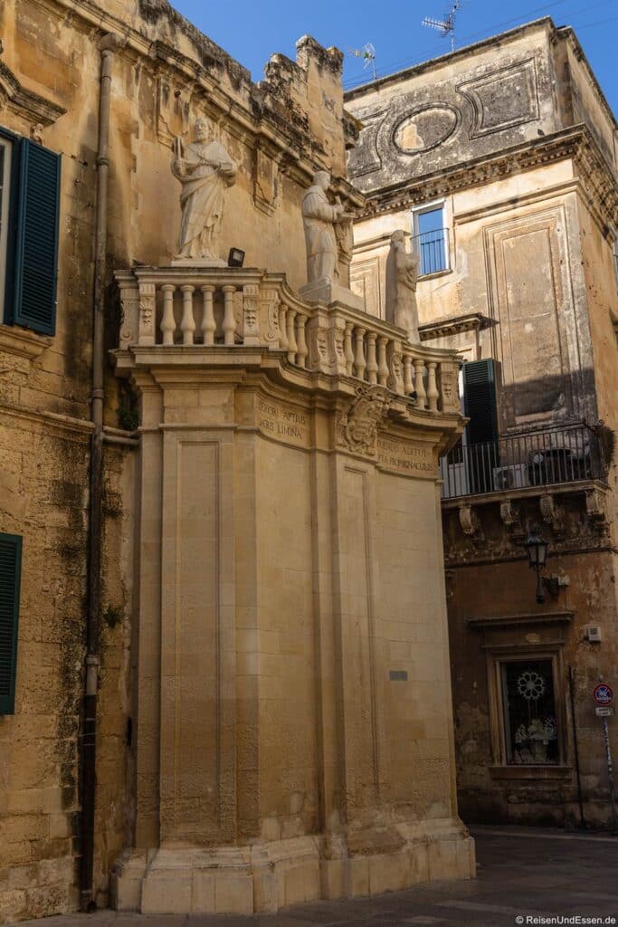 Lecce Sehenswürdigkeiten:Ein historisches Gebäude aus beigem Stein, auf dessen Eckgesims drei klassische Statuen thronen. Die Fassade des Gebäudes ist mit Säulen und lateinischen Inschriften verziert. Die angrenzenden Gebäude weisen verwitterten Stein, dunkle Fensterläden und Balkone vor einem blauen Himmel auf.