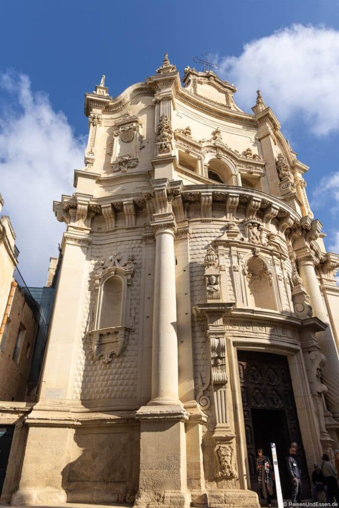 Kirchenfassade im Barockstil mit kunstvollem Mauerwerk, hohen Säulen, Bogenfenstern, dekorativen Schnitzereien und einer großen Holztür; das Gebäude liegt im Sonnenlicht vor einem teilweise bewölkten blauen Himmel.