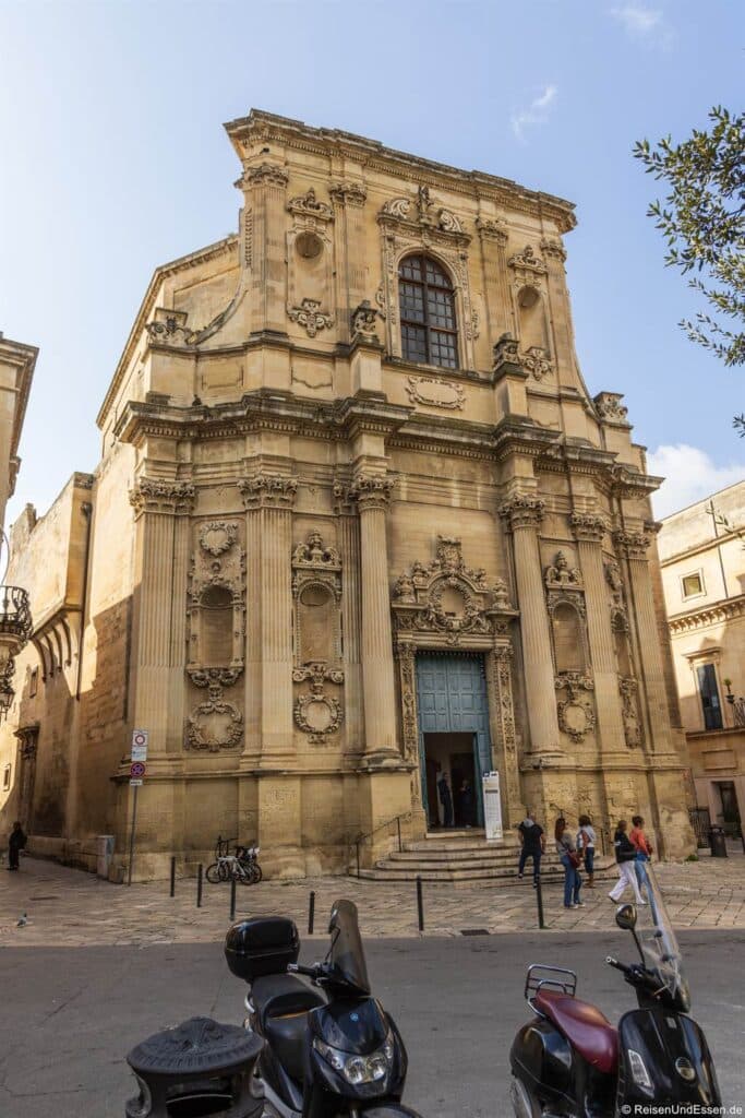 Lecce Sehenswürdigkeiten: Eine prächtige Barockfassade mit kunstvoll verzierten Säulen, Skulpturen und einem hohen Bogenfenster über einer blaugrünen Tür. Mehrere Menschen stehen und gehen in der Nähe des Eingangs. Im Vordergrund stehen Mopeds auf einer sonnigen, gepflasterten Straße.