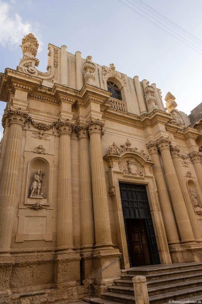 Lecce Sehenswürdigkeiten: Eine Kirchenfassade im Barockstil mit hohen Säulen, aufwendigen Steinmetzarbeiten, Statuen in Nischen, kunstvollen Verzierungen und einem großen Bogeneingang. Breite Steinstufen führen zum Eingang hinauf. Der Himmel ist teilweise bewölkt, und warmes Sonnenlicht lässt die Details des Gebäudes hervortreten.