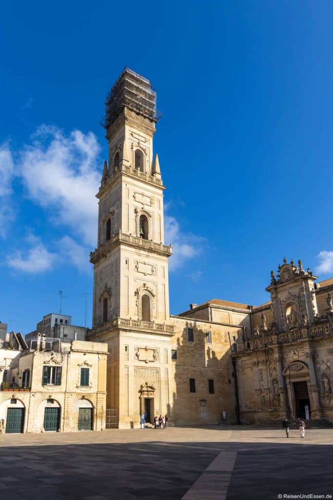 Ein hoher, historischer Glockenturm, dessen Spitze mit Gerüsten umgeben ist, steht auf einem sonnendurchfluteten Steinplatz neben kunstvollen Barockgebäuden unter einem strahlend blauen Himmel mit vereinzelten Wolken. Mehrere Menschen gehen um die Gebäude herum oder stehen in deren Nähe.