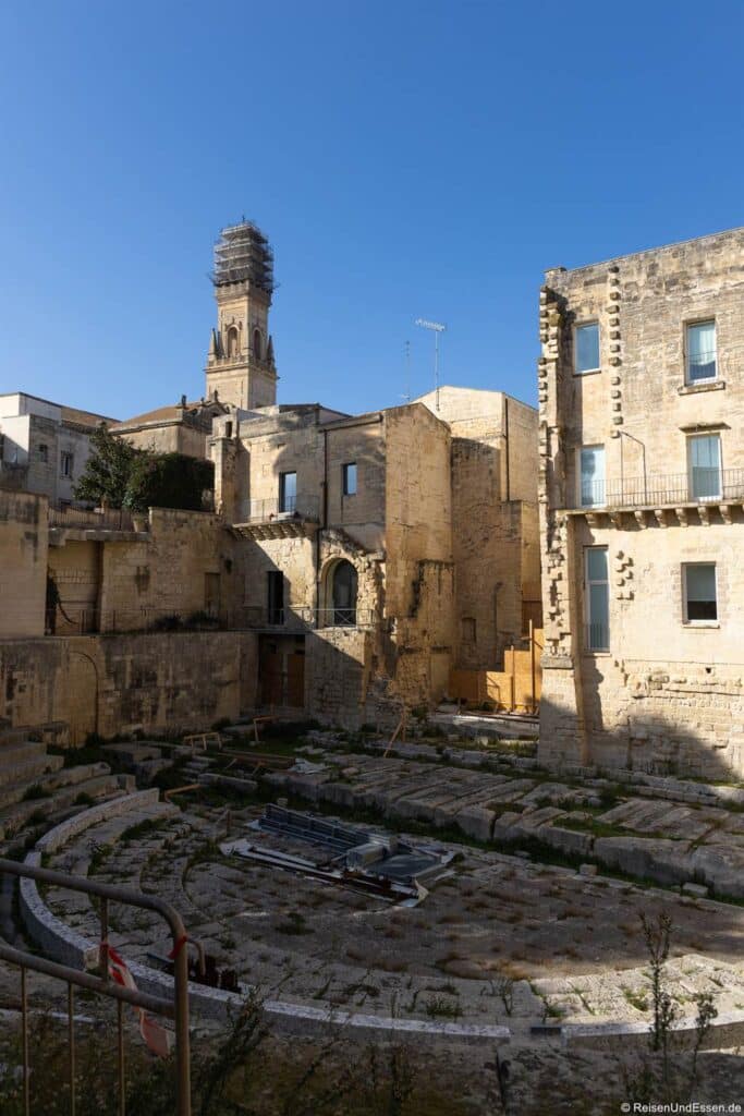 Ruinen eines antiken römischen Amphitheaters mit Reihen aus Steinsitzen, die mit Gras überwuchert sind und von alten, beigen Gebäuden umgeben sind. Im Hintergrund ist unter einem strahlend blauen Himmel ein hoher Glockenturm mit Gerüsten zu sehen.