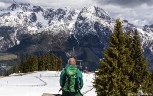 Eine Person mit einem grünen Rucksack mit der Aufschrift der Löwe sitzt vor einer Kulisse aus schneebedeckten Bergen und Kiefern. Ein Skilift verläuft schräg durch das Bild, und der bewölkte Himmel wirft Schatten auf die schroffen Gipfel. Der Boden ist mit Schnee bedeckt, dazwischen sind einige grüne Flecken zu sehen.