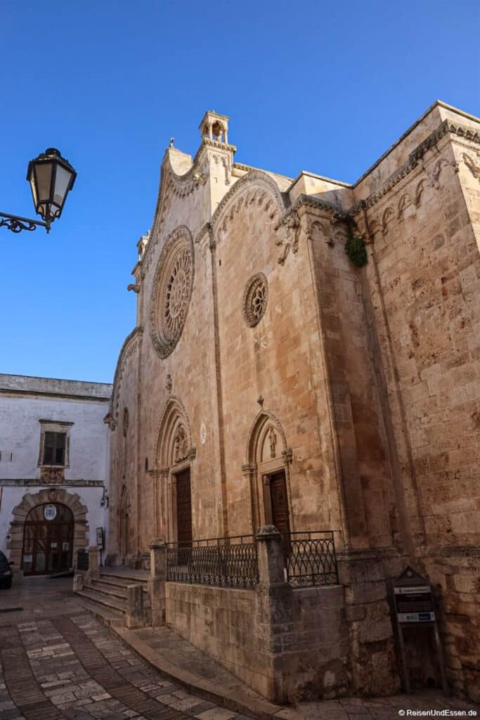 Ostuni Sehenswürdigkeiten: Unter einem strahlend blauen Himmel steht eine historische Steinkirche mit kunstvollen Schnitzereien, gewölbten Türen und einem großen Rosettenfenster. Das Gebäude verfügt über einen kleinen Glockenturm und liegt an einer Ecke einer Kopfsteinpflasterstraße, in deren Nähe sich eine Straßenlaterne und ein weiteres Gebäude befinden.