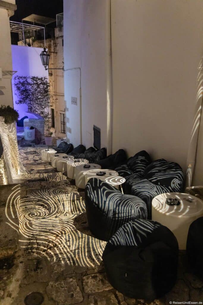 Eine schmale Steingasse bei Nacht, an deren Wänden runde, schwarze Stühle und weiße Tische aufgereiht sind. Kunstvolle Lichtmuster werden auf den Boden und die Möbel projiziert. Eine hohe weiße Wand und eine Straßenlaterne sind zu sehen und schaffen einen gemütlichen, modernen Außenbereich – eine einladende Ecke, die den Charme einfängt, den man oft bei den Sehenswürdigkeiten von Ostuni findet.