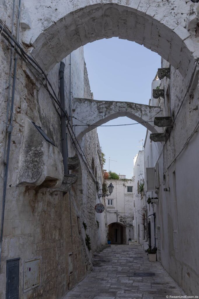 Ostuni Sehenswürdigkeiten: Eine schmale, gepflasterte Gasse in einer Altstadt mit weißen Mauern, Bogenbrücken über den Köpfen, einer rustikalen Laterne und geschlossenen Fensterläden. Die Straße ist leer, mit unebenen Steinen gepflastert, und an den Wänden verlaufen Kabel. Oben ist ein Stück Himmel und etwas Grün zu sehen.