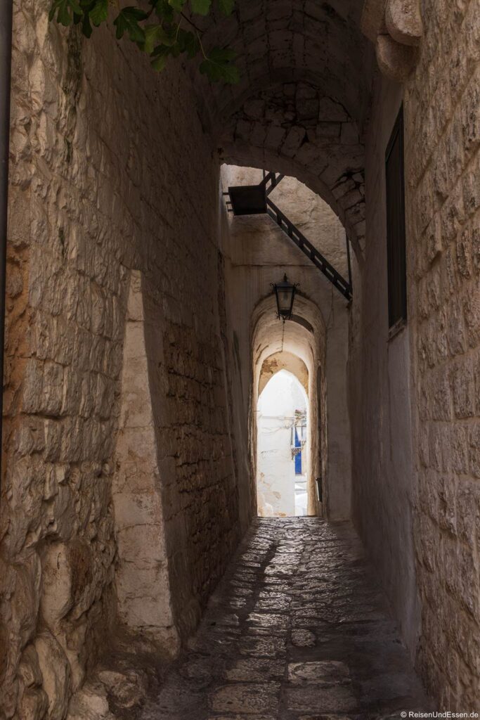 Eine schmale Steingasse mit gewölbter Decke, abgenutztem Kopfsteinpflaster und strukturierten Wänden; von der Decke hängt eine Laterne herab. Sonnenlicht strömt durch einen offenen Durchgang am Ende der Gasse und gibt den Blick auf weiße Gebäude und den blauen Himmel draußen frei.