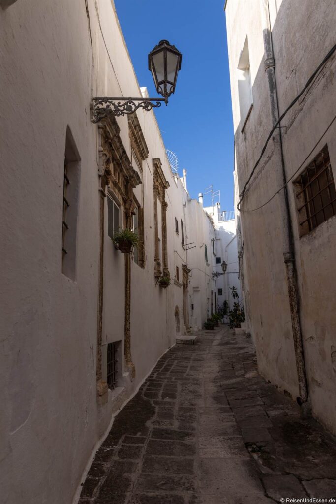 Ostuni Sehenswürdigkeiten: Eine schmale Kopfsteinpflastergasse mit weiß getünchten Gebäuden, verzierten Fensterrahmen, einer hängenden Laterne und blauem Himmel darüber. Der Weg macht eine leichte Kurve, am Ende sind Topfpflanzen zu sehen, was eine ruhige mediterrane Atmosphäre schafft, die an die Sehenswürdigkeiten von Ostuni erinnert.