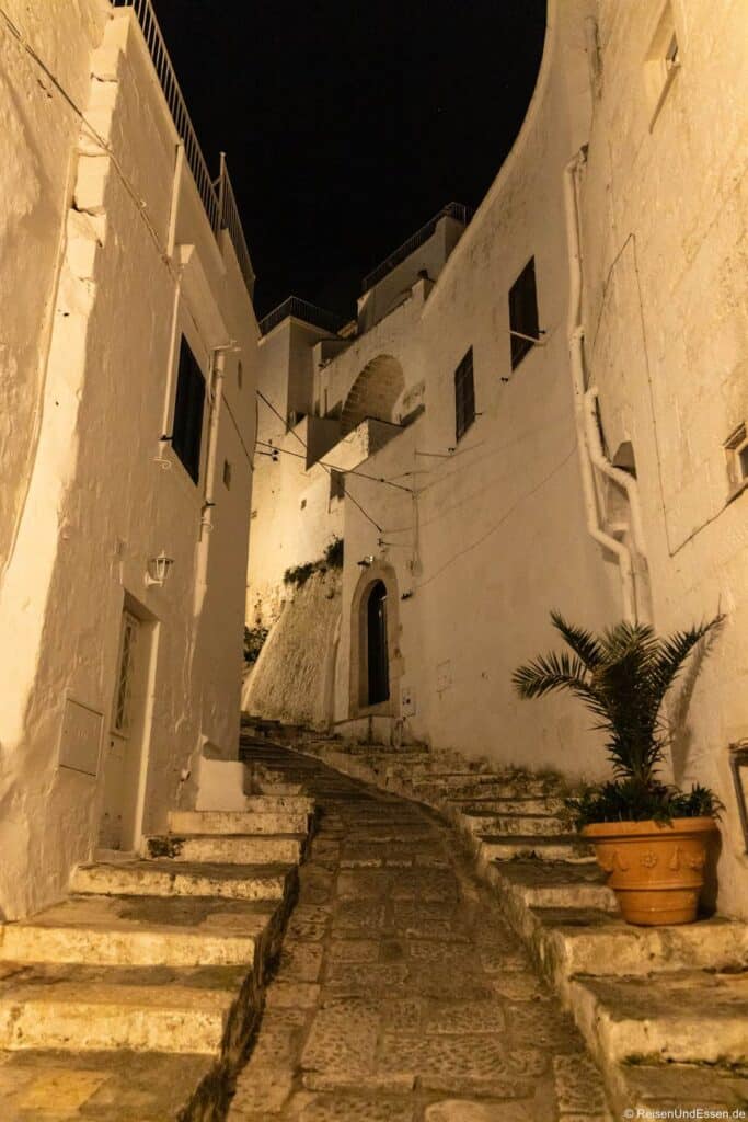 Eine schmale, ruhige Steingasse in einem mediterranen Dorf bei Nacht, gesäumt von hohen, weiß getünchten Gebäuden mit gewölbten Türöffnungen – die an die Sehenswürdigkeiten von Ostuni erinnern. Entlang des Weges befinden sich Stufen und rechts steht eine Palme in einem Topf. Warmes gelbes Licht hebt die strukturierten Wände und den Weg hervor.