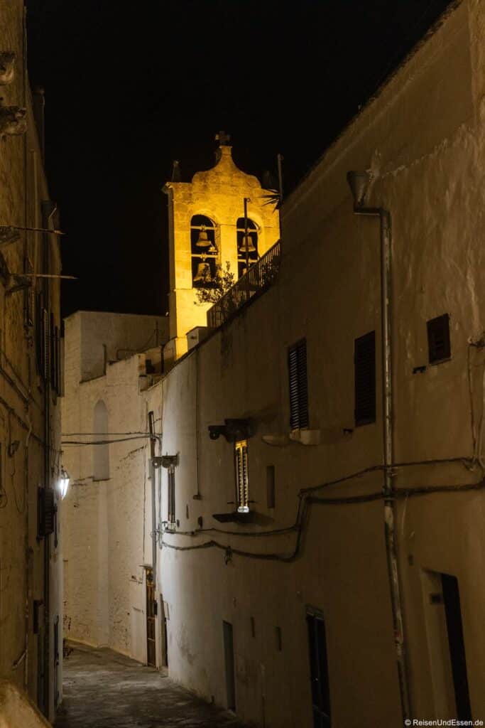 Eine schmale, schummrig beleuchtete Gasse mit weißen Wänden führt nachts bergauf. Im Hintergrund leuchtet ein Glockenturm im gelben Schein der Scheinwerfer. An den Wänden sind Fenster und Stromleitungen zu sehen. Der Himmel ist völlig dunkel, was den beleuchteten Turm besonders hervorhebt.