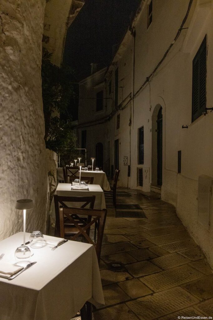 Eine schmale Kopfsteinpflastergasse bei Nacht mit weißen Wänden. Drei kleine Tische sind mit weißen Tischdecken, Gläsern, Besteck und Lampen gedeckt und schaffen eine gemütliche, intime Atmosphäre für ein Essen im Freien. Die Szene ist ruhig und wird von den Tischlampen warm beleuchtet – sie fängt den authentischen Charme der Sehenswürdigkeiten von Ostuni ein.