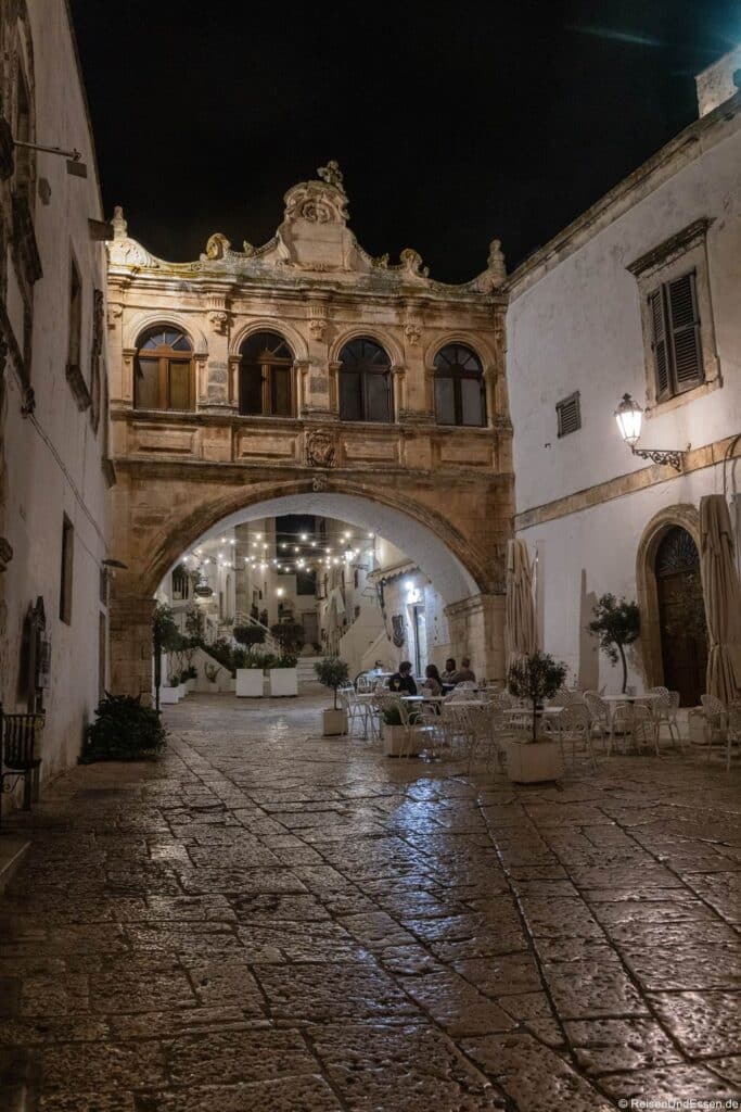 Eine nächtliche Ansicht eines historischen europäischen Steinbogenportals mit kunstvollen Details, beleuchtet von warmen Straßenlaternen. Darunter stehen leere weiße Tische und Stühle auf einer glänzenden Kopfsteinpflasterstraße. Im Hintergrund sind Menschen in der Nähe weißer Gebäude zu sehen.