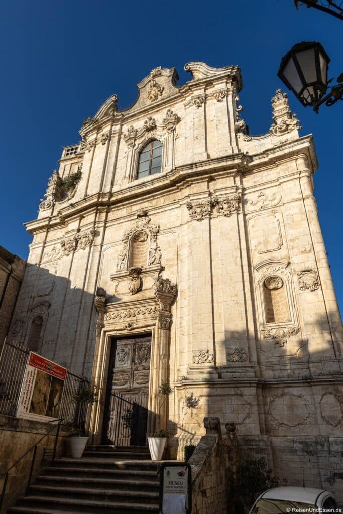 Ostuni Sehenswürdigkeiten: Eine Kirche im Barockstil mit einer reich verzierten Steinfassade, einem Bogenportal, aufwendigen Schnitzereien, drei Bogenfenstern und dekorativen Reliefs. Eine breite Steintreppe führt zum Eingang – ein klassisches Beispiel unter den Sehenswürdigkeiten von Ostuni. Rechts hängt eine Straßenlaterne. Helles Sonnenlicht wirft tiefe Schatten unter einem strahlend blauen Himmel.