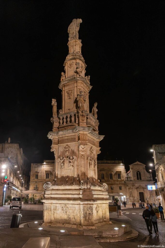 Ein hohes, kunstvoll verziertes Steinmonument mit zahlreichen Statuen und Reliefs, das nachts auf einem Stadtplatz beleuchtet wird. Historische Gebäude mit Bogenfenstern umgeben das Monument. Unter dem dunklen Himmel sind nur wenige Menschen in der Nähe unterwegs.