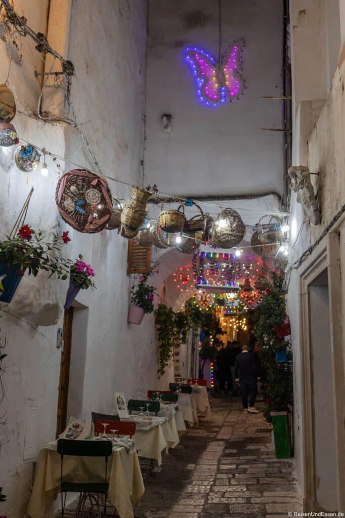 Eine schmale, mit Kopfsteinpflaster gepflasterte Gasse mit kleinen Tischen und Stühlen, Hängekörben und Pflanzen rundet das Bild ab. Über ihnen hängen bunte Lichter und ein leuchtend violetter Schmetterling. Am anderen Ende versammeln sich Menschen und sorgen für eine gemütliche, festliche Atmosphäre.