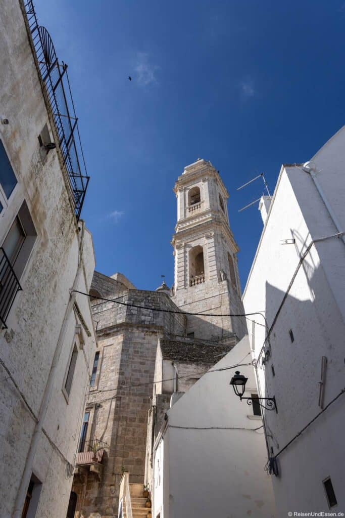 Eine schmale Straße mit weiß getünchten Häusern führt unter einem strahlend blauen Himmel zu einem hohen Glockenturm aus Stein. Die Schatten der Gebäude und einer schwarzen Laterne sind zu sehen und bilden einen Kontrast zwischen hellem Sonnenlicht und schattigen Wänden.