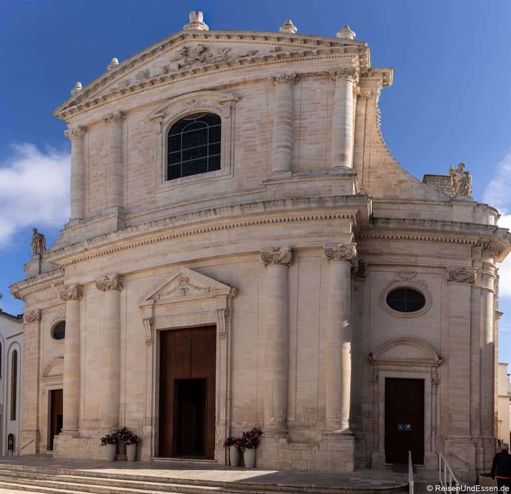 Eine imposante beigefarbene Steinkirche, eine der bemerkenswertesten Sehenswürdigkeiten in Locorotondo, besticht durch klassische Säulen, kunstvolle Skulpturen, Bogenfenster und drei große Holztüren. Eine Treppe führt zum Eingang hinauf, der von Blumentöpfen flankiert wird. Der Himmel ist strahlend blau und mit vereinzelten Wolken übersät.