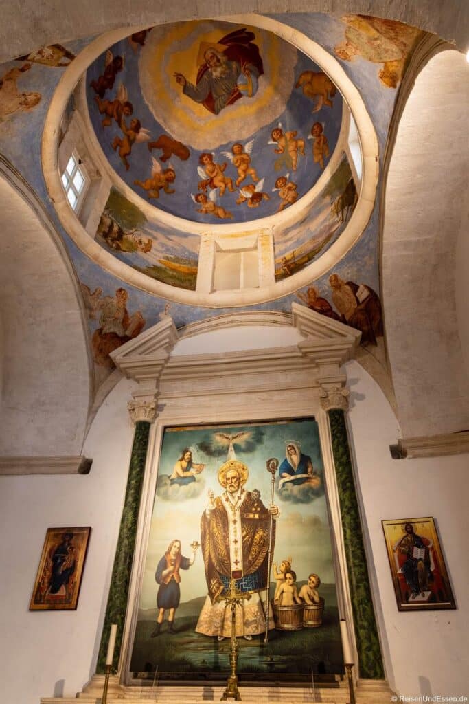 Innenraum einer Kirche mit Kuppeldecke, an der ein Fresko zu sehen ist, das Gott inmitten von Engeln darstellt. Darunter befindet sich ein großes Gemälde eines Heiligen mit einem Stab, das von zwei grünen Säulen flankiert wird und zwischen zwei kleineren, gerahmten religiösen Darstellungen an den weißen Wänden zentriert ist.