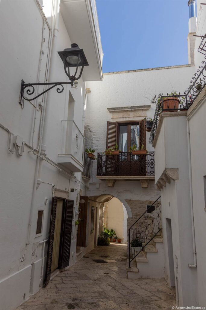 Eine schmale Gasse mit weiß getünchten Wänden, einem schmiedeeisernen Balkon mit Pflanzen, Fenstern mit Fensterläden, einer Steintreppe mit Geländer und einem gewölbten Durchgang. Über dem Ganzen ist eine schwarze Laterne an der Wand befestigt. Helles Sonnenlicht taucht die Szene in helles Licht.