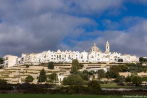 Das italienische Bergdorf Locarotondo mit weißen Steinhäusern und einer großen Kirche mit hohem Glockenturm vor einem teilweise bewölkten blauen Himmel. Terrassengärten und Bäume bedecken den Hang im Vordergrund.