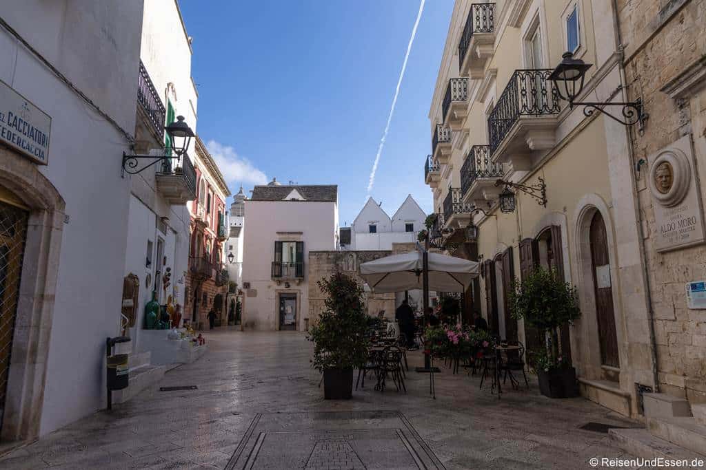 Locorotondo Sehenswürdigkeiten: Eine ruhige, sonnendurchflutete Straße in einer europäischen Stadt mit Kopfsteinpflaster, historischen Gebäuden, Balkonen und einem Café mit Sitzplätzen im Freien unter Sonnenschirmen versetzt den Betrachter in den Charme vieler Sehenswürdigkeiten in Locorotondo. Blumentöpfe und Straßenlaternen säumen die einladende Straße; der Himmel ist blau, und über ihr zieht eine dünne weiße Kondensstreifen.