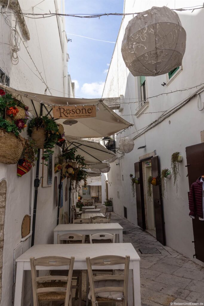 Eine schmale Kopfsteinpflastergasse mit weißen Wänden, hängenden Blumenkörben, zwei weißen Tischen mit Stühlen, Sonnenschirmen mit der Aufschrift „Ristorante“, Lichterketten und großen dekorativen Laternen über den Köpfen. An der Gasse reihen sich Türen und Fenster aneinander; der Himmel ist teilweise bewölkt.