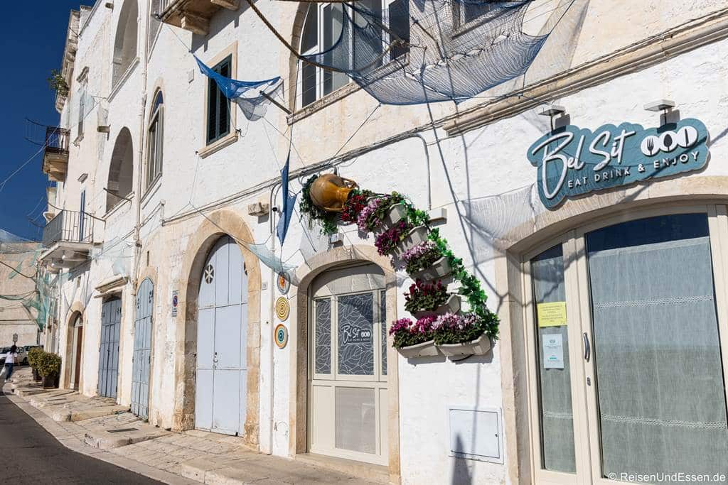Ein weißes Gebäude mit gewölbten Türen und Fenstern, über denen Fischernetze hängen, und einem Schild mit der Aufschrift „BelSit Eat Drink & Enjoy“. An der Wand hängt ein großer Krug, der mit rosa und weißen Blumen verziert ist. Helles Sonnenlicht wirft Schatten auf die gepflasterte Straße.