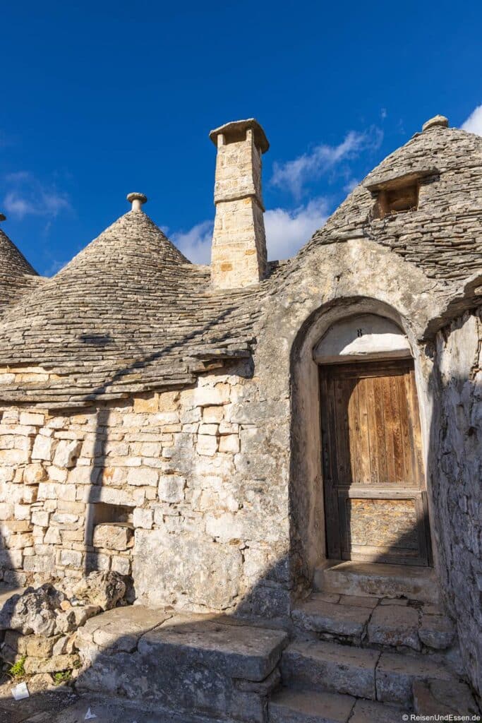 Ein Trullo-Haus aus Stein in Alberobello zeichnet sich durch ein kegelförmiges Dach aus aufgeschichteten Steinen und eine verwitterte Holztür aus. Ein hoher Steinkamin ragt vor dem strahlend blauen Himmel empor, während die Wände aus grobem Mauerwerk im hellen Sonnenlicht scharfe Schatten werfen.
