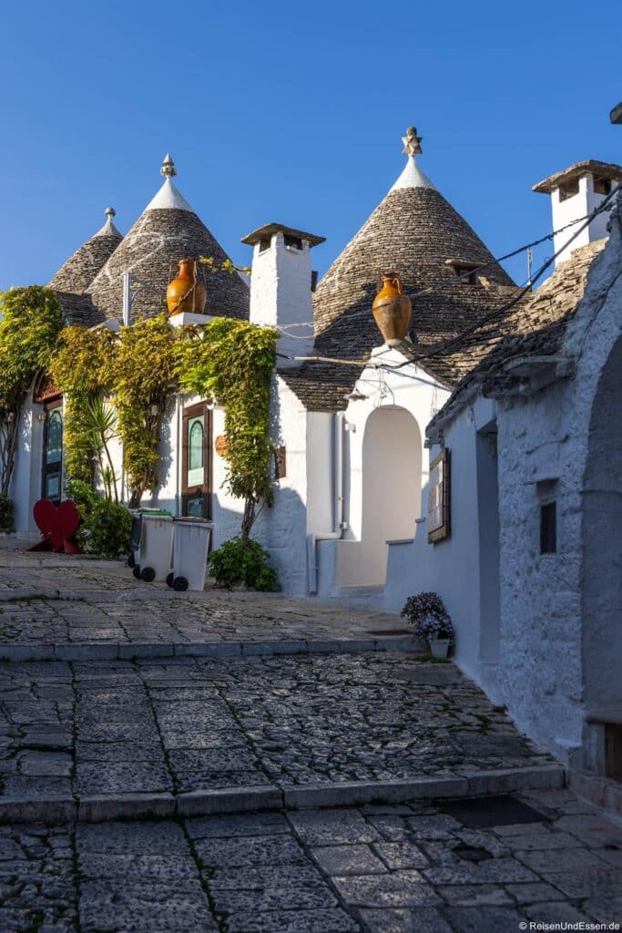 Traditionelle weiß getünchte Trulli-Häuser mit kegelförmigen Steindächern säumen eine Kopfsteinpflasterstraße unter einem strahlend blauen Himmel. Die Gebäude sind mit Kletterpflanzen, Terrakotta-Vasen und einer kleinen roten Herzskulptur neben einer weißen Mülltonne geschmückt.