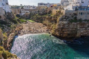 Ein kleiner Sandstrand liegt eingebettet zwischen steilen, felsigen Klippen und klarem, türkisfarbenem Wasser. Die Menschen entspannen sich im Sand und schwimmen im Meer, während weiße Gebäude und eine Steinbrücke die Klippenkrone säumen und die Bucht überragen.