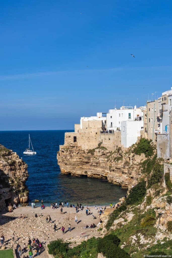 Ein kleiner Sandstrand schmiegt sich zwischen felsigen Klippen unterhalb einer Reihe weißer mediterraner Gebäude ein. Ein einsames Segelboot treibt unter klarem Himmel auf dem tiefblauen Meer, während unten am Ufer Menschen spazieren gehen.