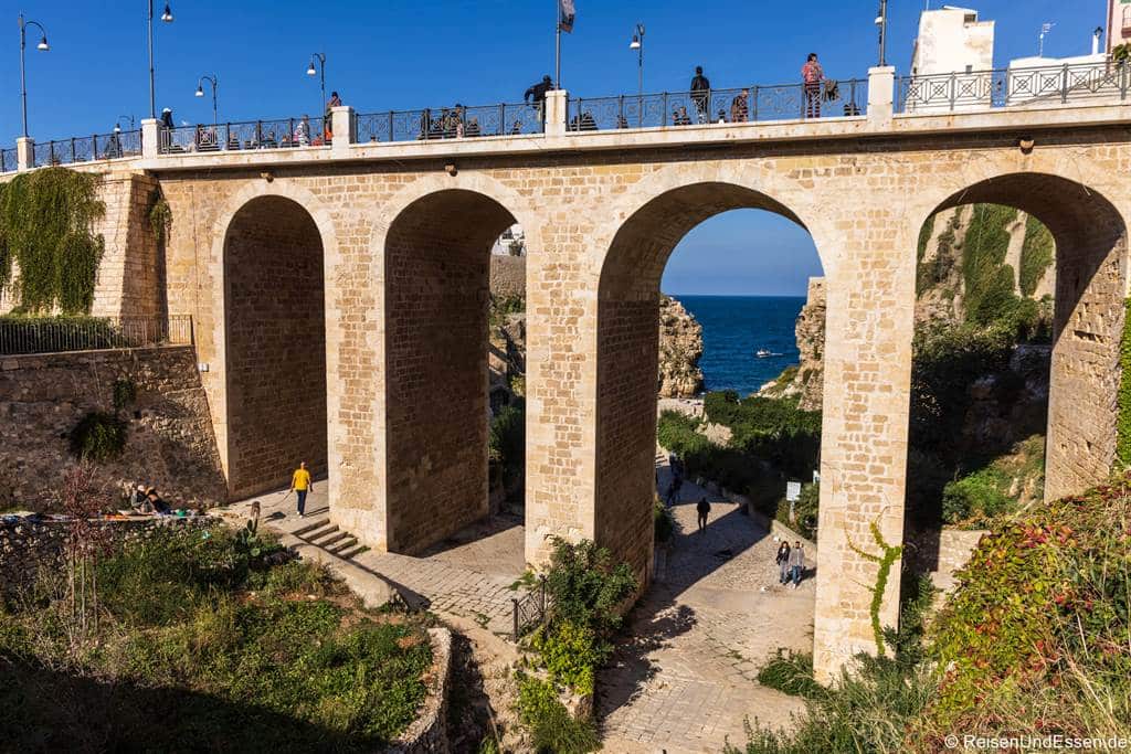 Eine große Steinbrücke mit vier Bögen überspannt eine Schlucht; Menschen spazieren auf der Brückendecke und durch die Schatten darunter. Der Blick durch den mittleren Bogen offenbart das strahlend blaue Meer, während üppig grüne Pflanzen am verwitterten Brückenfuß emporwachsen.