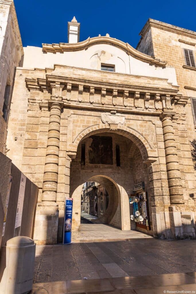 Ein steinerner Torbogen in Polignano a Mare bildet den Eingang zu einer schmalen Stadtstraße unter strahlend blauem Himmel. Das historische Tor ist mit verzierten Säulen und einem Wappen in der Mitte geschmückt; an der Seite des Bogens befindet sich das Schaufenster eines kleinen Ladens.