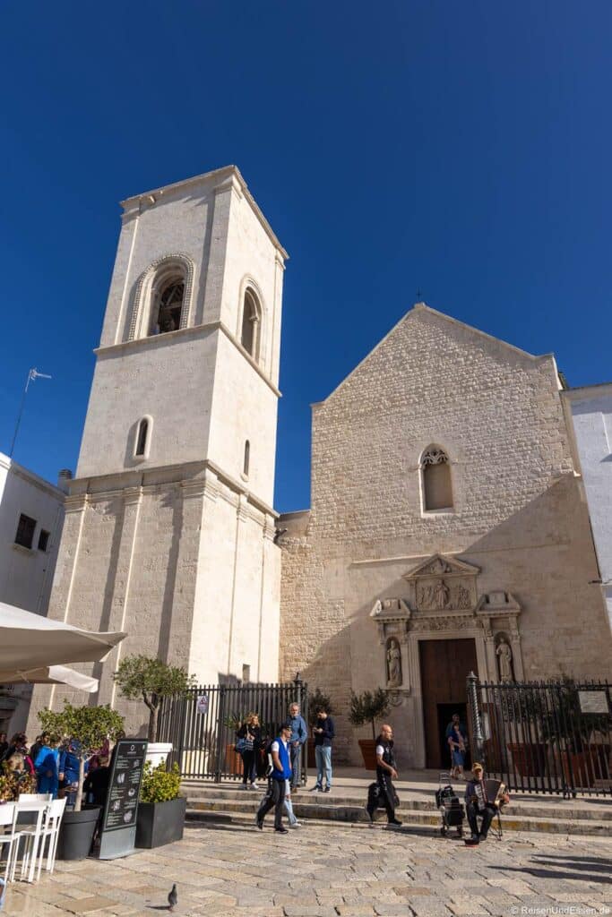 Polignano a Mare Ausflug: Unter einem strahlend blauen Himmel steht eine hohe romanische Steinkirche. Neben der Giebel-Fassade erhebt sich ein quadratischer Glockenturm, während im Vordergrund Menschen über einen sonnigen Steinplatz schlendern.