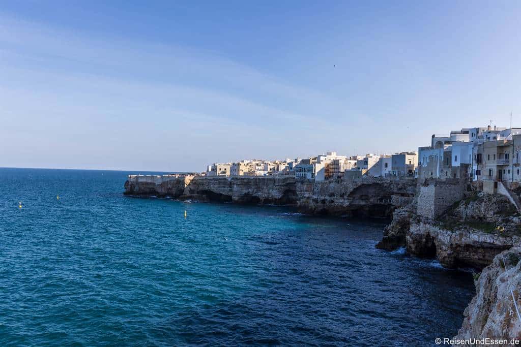 Die Küstenstadt Polignano a Mare thront auf hohen, zerklüfteten Kalksteinfelsen mit Blick auf die tiefblaue Adria. Unter einem klaren, strahlend blauen Himmel reihen sich weiß getünchte Gebäude am Rand der Klippen aneinander.
