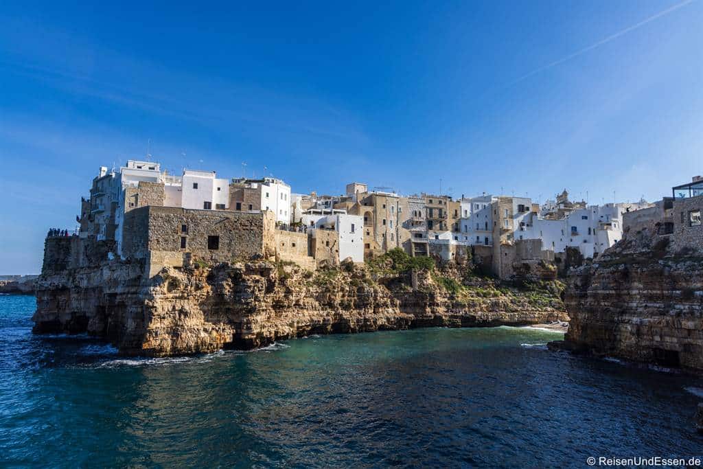 Polignano a Mare Ausflug: Eine mediterrane Küstenstadt wie Polignano a Mare liegt auf schroffen Klippen mit Blick auf das blaue Meer. Die Gebäude sind überwiegend weiß und haben Sockel aus Naturstein, die einen schönen Kontrast zum klaren, tiefblauen Himmel bilden.