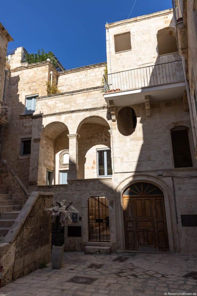 Ein sonnendurchfluteter Innenhof in einem Gebäudekomplex aus Stein besticht durch Bogenöffnungen, einen kleinen Balkon und einen hölzernen Bogenportal. Die verwitterten, hellen Steinmauern werfen unter einem strahlend blauen Himmel lange Schatten.