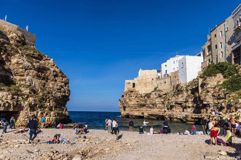 In einer Bucht zwischen hohen Kalksteinfelsen, auf denen weiße Gebäude thronen, liegt ein Kieselstrand. Menschen in Freizeitkleidung entspannen sich auf den Steinen unter einem klaren, strahlend blauen Himmel.