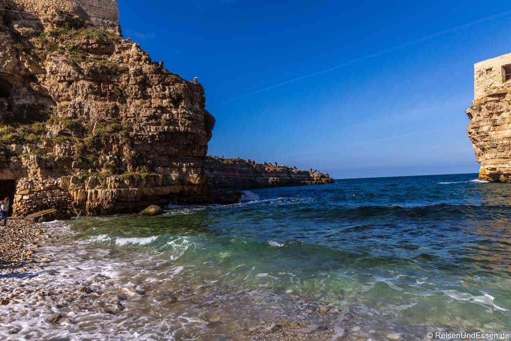 Zwischen zwei hoch aufragenden, verwitterten Steinfelsen am Ufer eines tiefblauen Meeres erstreckt sich ein Kieselstrand. Sanfte Wellen plätschern unter einem klaren, strahlenden Himmel an den Strand.