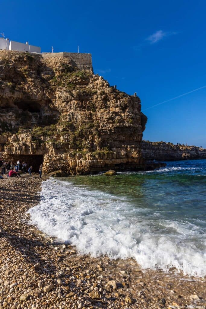 Polignano a Mare Ausflug: Ein Kieselstrand trifft auf das klare, türkisfarbene Meer unterhalb einer hoch aufragenden, schroffen Felswand. Kleine Gestalten stehen in der Nähe eines Höhleneingangs am Fuße des Felsens, während sich ein weißes Gebäude auf der Klippe vor einem tiefblauen Himmel abhebt.