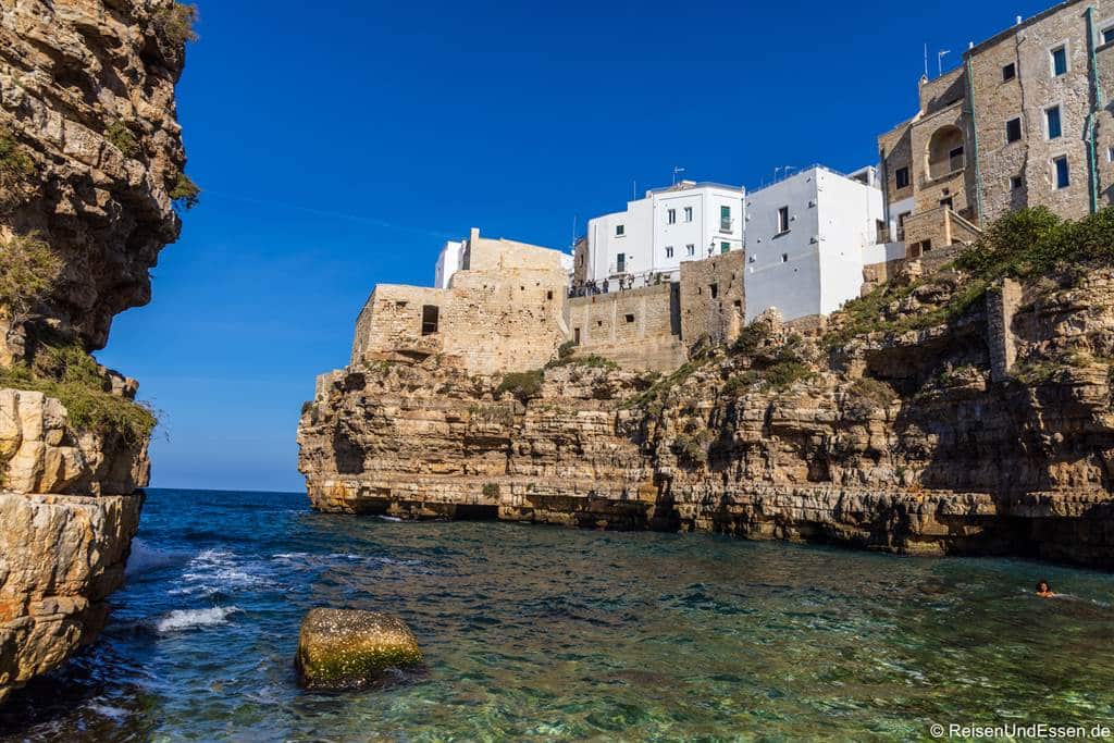 Weiße Gebäude thronen auf zerklüfteten, felsigen Klippen mit Blick auf ein klares, blaues Meer. Eine schmale Bucht trennt die Felsformationen voneinander, und rechts im Wasser ist ein Schwimmer zu sehen. Der Himmel ist tiefblau und wolkenlos.