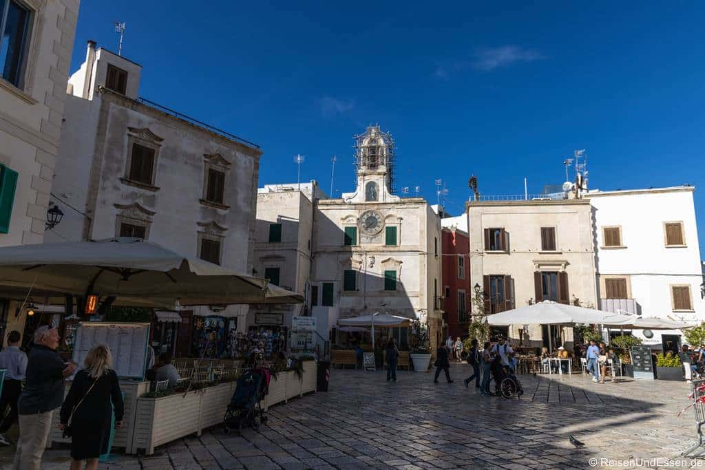 Polignano a Mare Ausflug: Ein sonniger Steinplatz in einer italienischen Stadt ist von historischen Gebäuden und Straßencafés umgeben. Über der zentralen Fassade thront ein Uhrturm, während Menschen unter strahlend blauem Himmel über den Platz schlendern.