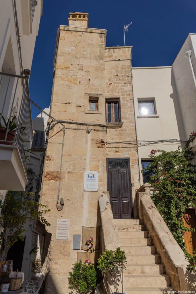 Am oberen Ende einer kurzen Steintreppe steht ein hohes, hellbraunes Steingebäude mit einer dunklen Holztür. An der Fassade befinden sich ein kleines Schild, eine Wandtafel und hängende Zierelemente, die sich alle vor einem klaren, tiefblauen Himmel abheben.